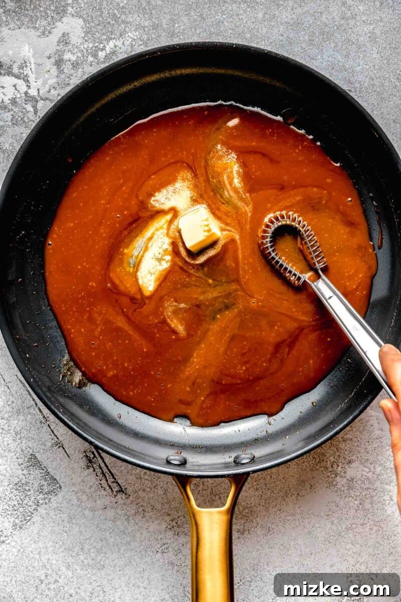 The crucial step of enriching the bourbon sauce by gradually incorporating chilled butter cubes. Adding small cubes of chilled unsalted butter to a simmering sauce in a pan.