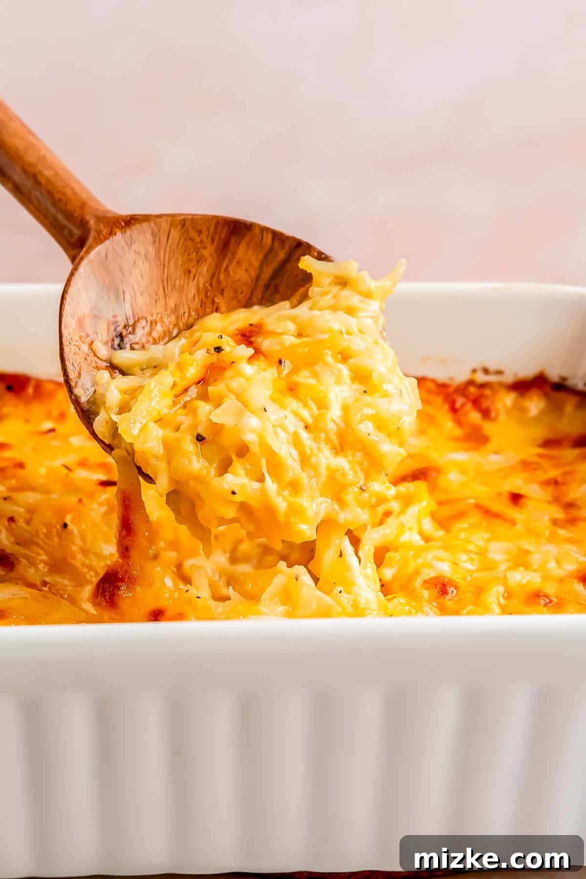 Irresistible Cheesy Hashbrown Bake 3 A wooden spoon scoops a generous portion of warm, gooey cheesy hashbrown casserole from a white baking dish.