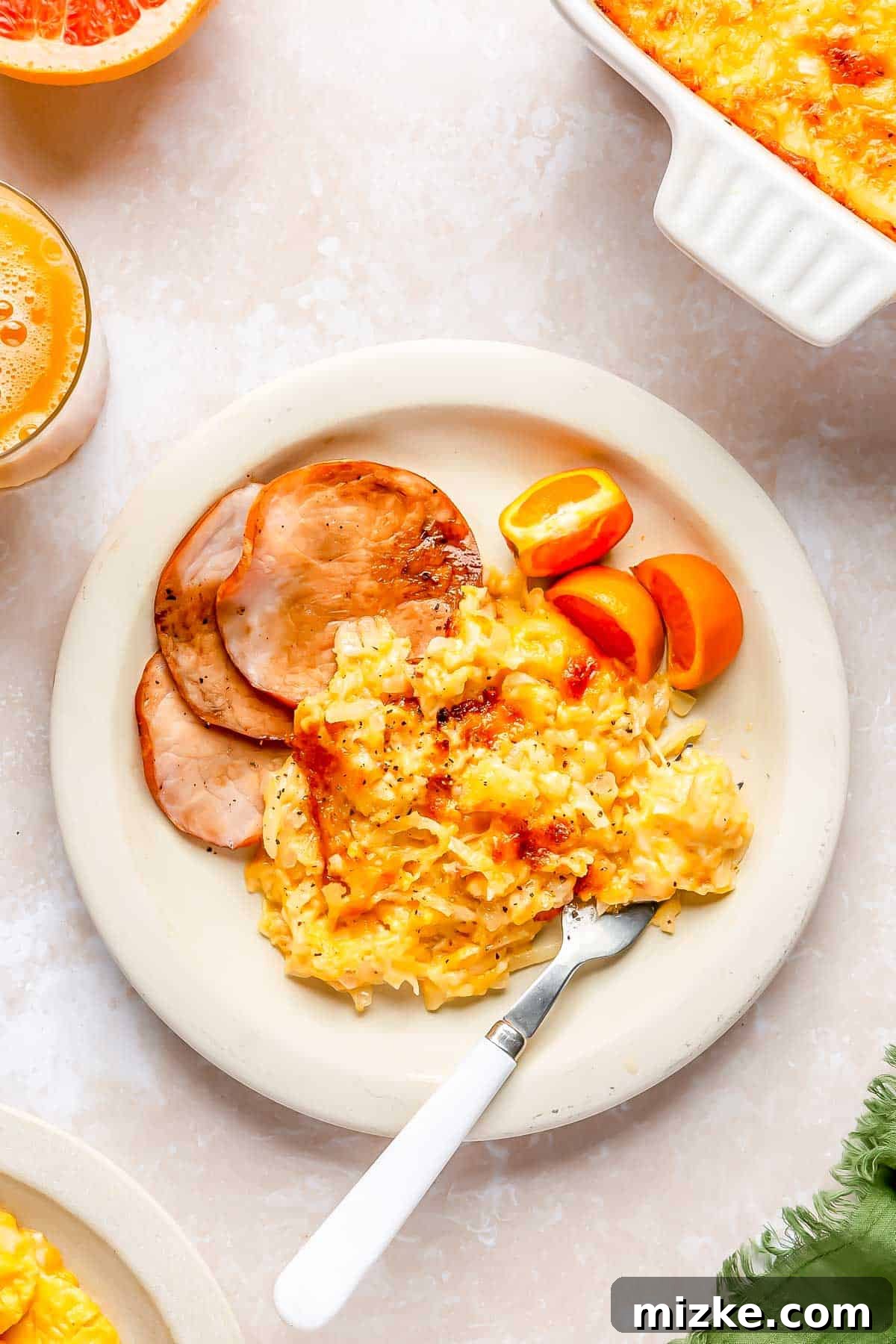 Irresistible Cheesy Hashbrown Bake 4 A plate featuring a generous serving of cheesy hashbrown casserole, alongside slices of Canadian bacon and fresh orange slices, creating a complete breakfast.
