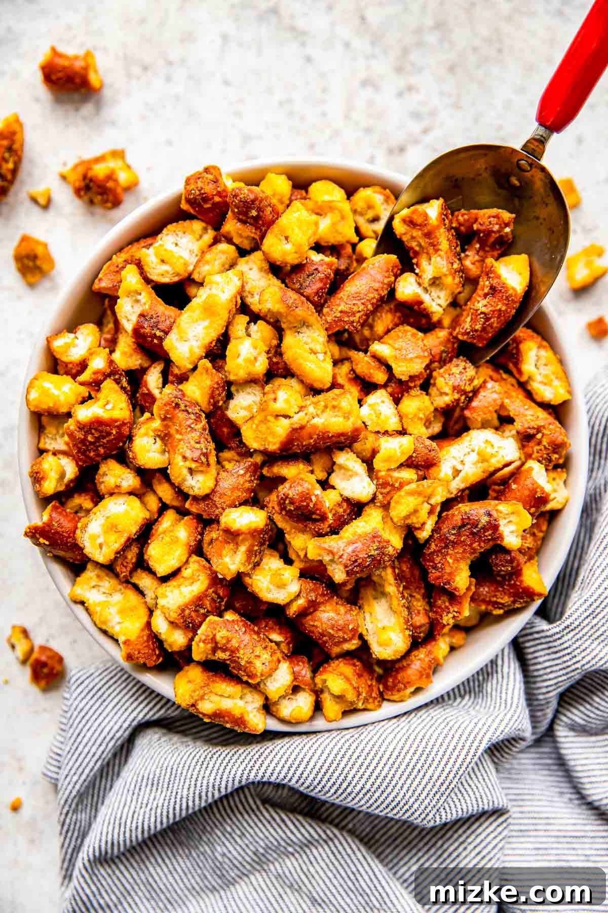 Tangy Gold Pretzel Crunch 2 A scoop delves into a generously filled bowl of golden-brown homemade honey mustard pretzels, coated in a delightful seasoning.