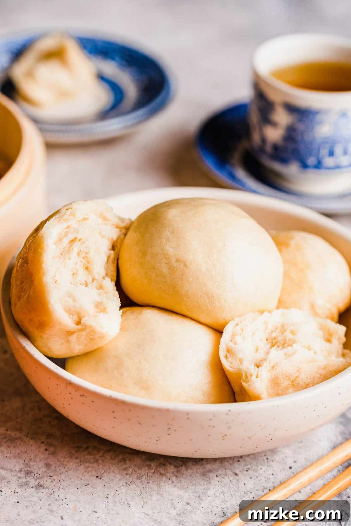 These Chinese steamed buns are moist, fluffy, sweet, and only use 5 ingredients. They're perfect on their own, or as an Asian side dish. A bowl full of Chinese steamed buns, including one that's torn in half, with a cup of tea in the background
