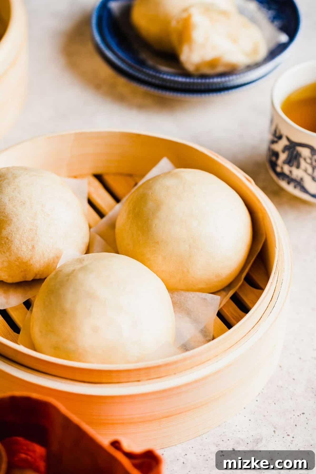 These Chinese steamed buns are moist, fluffy, sweet, and only use 5 ingredients. They're perfect on their own, or as an Asian side dish. Three Chinese steamed buns sitting in a steamer, with a bun on a plate in the background, next to a cup of tea