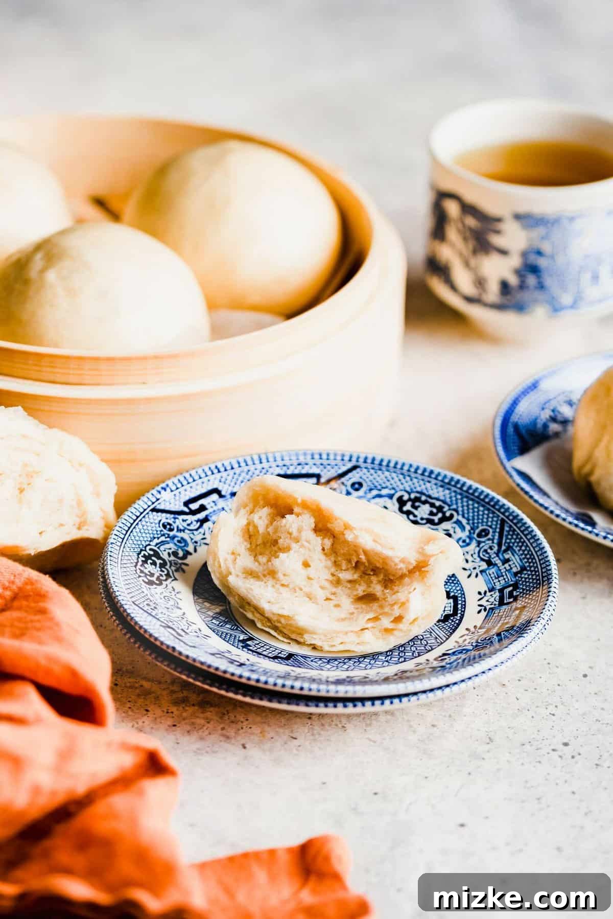 These Chinese steamed buns are moist, fluffy, sweet, and only use 5 ingredients. They're perfect on their own, or as an Asian side dish. Half of a Chinese steamed bun on a plate, next to another plate, with a steamer full of buns and a cup of tea in the background