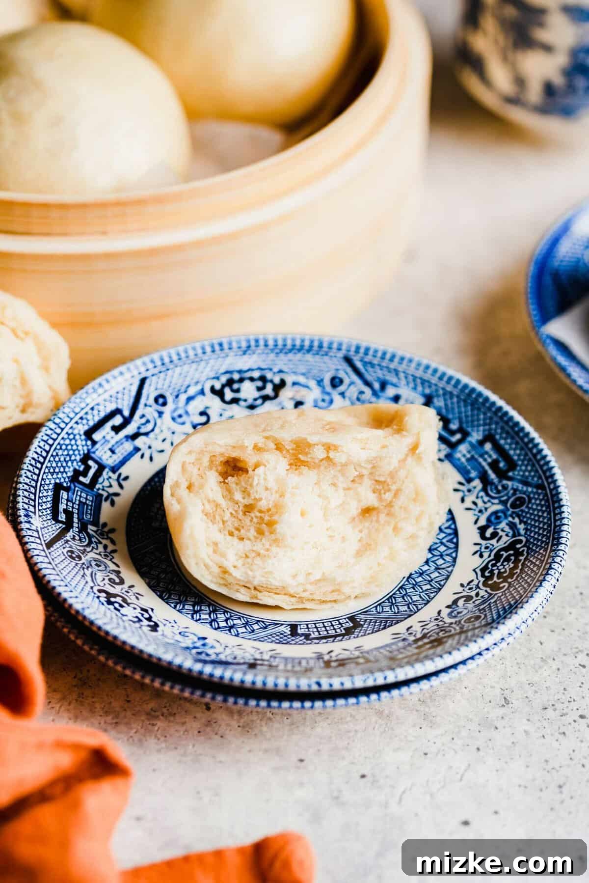 These Chinese steamed buns are moist, fluffy, sweet, and only use 5 ingredients. They're perfect on their own, or as an Asian side dish. A half-eaten Chinese steamed bun on a plate, with a steamer full of buns in the background
