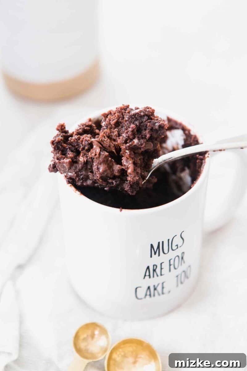 The moistest chocolate mug cake you will ever have! It's not spongy like other mug cakes! Recipe on tablefortwoblog.com A large spoonful of microwaved chocolate mug cake sitting balancing on top of a white mug that says "Mugs Are For Cakes, Too" and gold measuring spoons at the bottom of the image.
