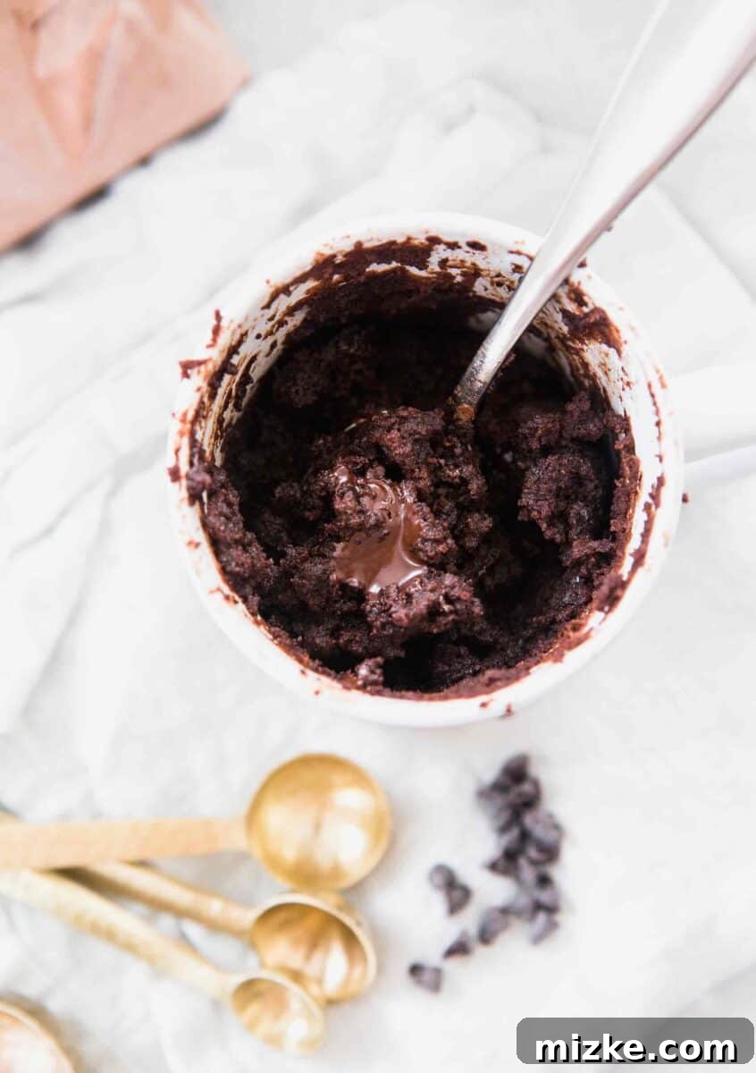 The moistest chocolate mug cake you will ever have! It's not spongy like other mug cakes! Recipe on tablefortwoblog.com An overhead shot of the chocolate mug cake with hazelnut spread in the center. The mini chocolate chips and gold measuring spoons are on the side of the mug cake and sitting on a white linen towel.