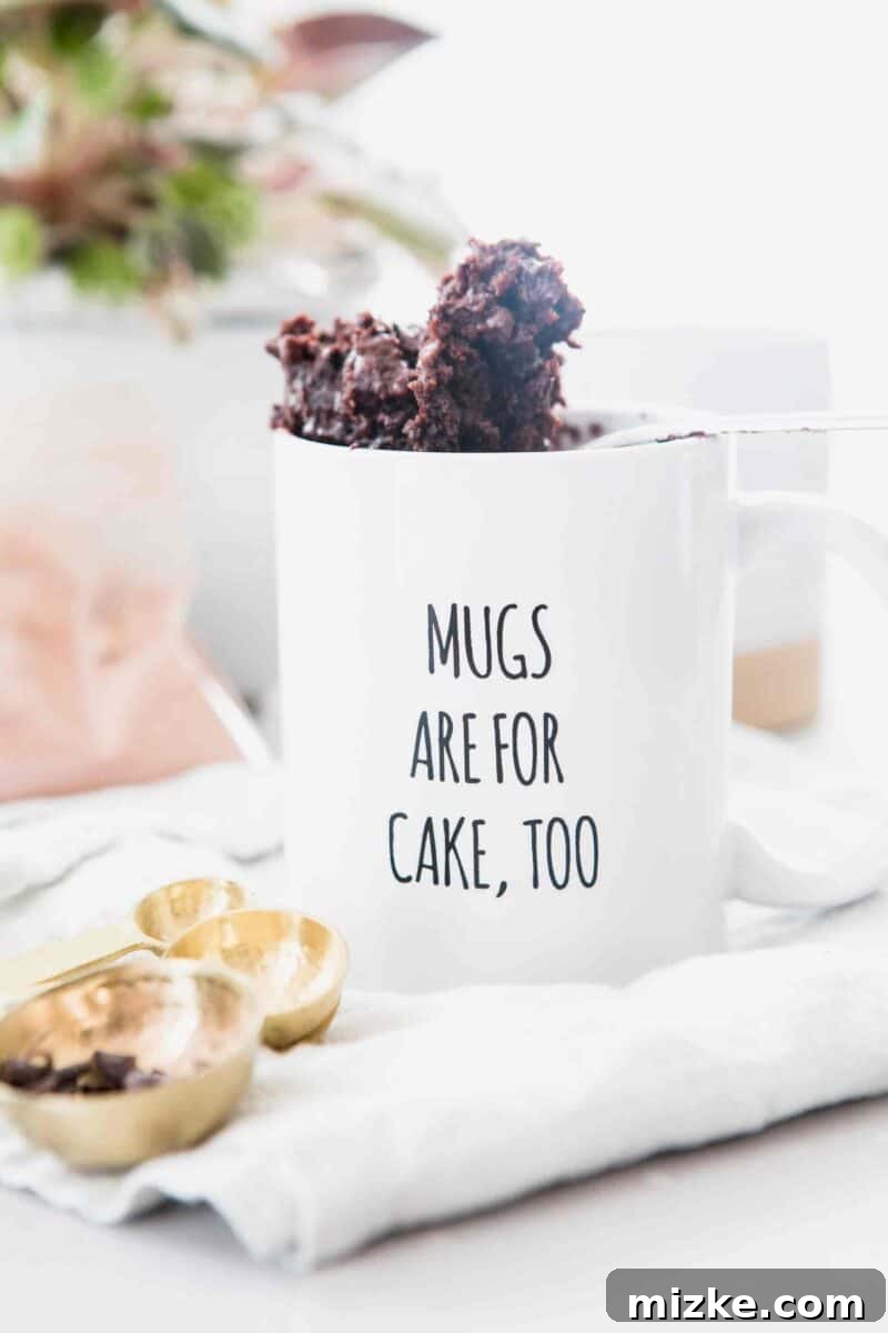 The moistest chocolate mug cake you will ever have! It's not spongy like other mug cakes! Recipe on tablefortwoblog.com A spoonful of chocolate mug cake peeking out of a white mug that says "Mugs Are For Cake, Too" on top of a white linen towel with a measuring spoon of mini chocolate chips.