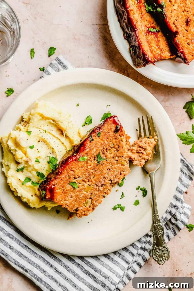 Enjoy a comforting plate of classic meatloaf paired with fluffy mashed potatoes – a truly satisfying weeknight meal. A perfectly sliced portion of classic meatloaf served on a plate with creamy mashed potatoes and green garnish.