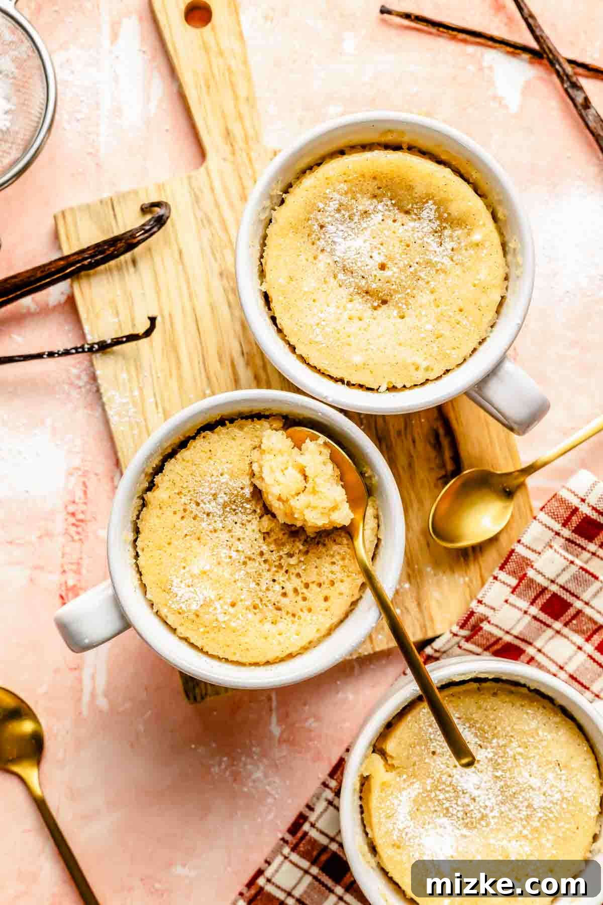 Pure Vanilla Mug Cake 2 Three vanilla mug cakes on a wooden cutting board with a gold spoon digging out the center of one mug cake, highlighting its fluffy texture.