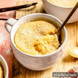 A golden spoon has scooped out a quarter of the vanilla cake in the mug, showcasing its golden-brown top and soft, moist interior.