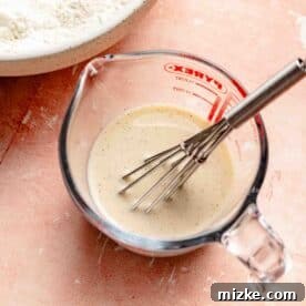 Milk mixture dotted with vanilla beans in a glass measuring cup with a metal whisk, showing the liquid ingredients.