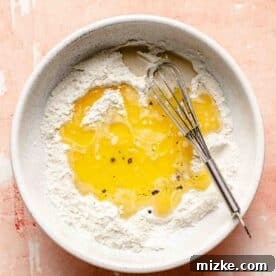 Melted butter and liquid milk mixture added to a small bowl of flour mixture with a metal whisk in the center, ready for mixing.