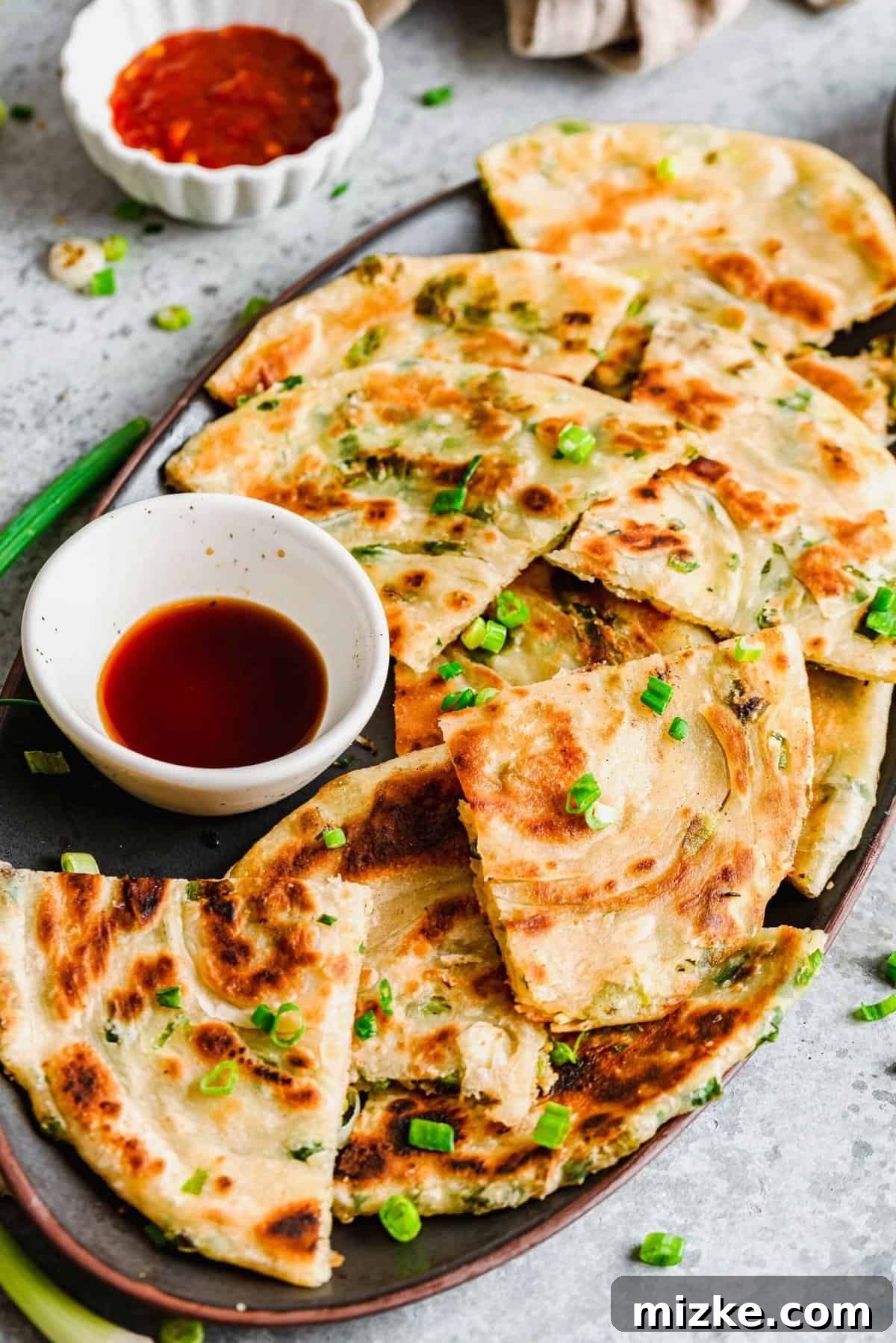 Homemade scallion pancakes are crisp on the outside, delightfully chewy on the inside, and easier to make than you think! Learn how to create these flaky layers at home. Golden-brown scallion pancakes cut into triangles, arranged beautifully on a serving plate with a side of dipping sauce.