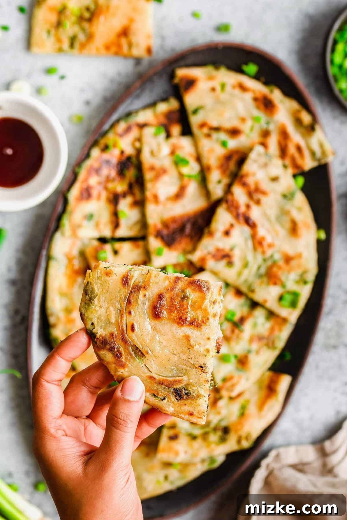Experience the delightful crispness and chewiness of these homemade scallion pancakes. Making them at home is easier than you might think! A hand holding a perfectly cooked scallion pancake, showcasing its golden-brown crispness, above a platter of more pancakes ready for serving.