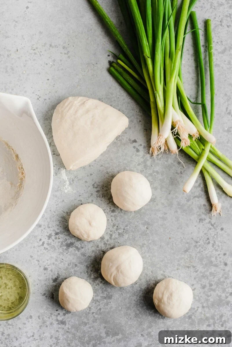 Portioned dough balls, fresh scallions, and oil: all the simple ingredients ready for assembling delicious homemade scallion pancakes. Individual balls of scallion pancake dough neatly arranged on a work surface, accompanied by finely chopped scallions and a bowl of vegetable oil.