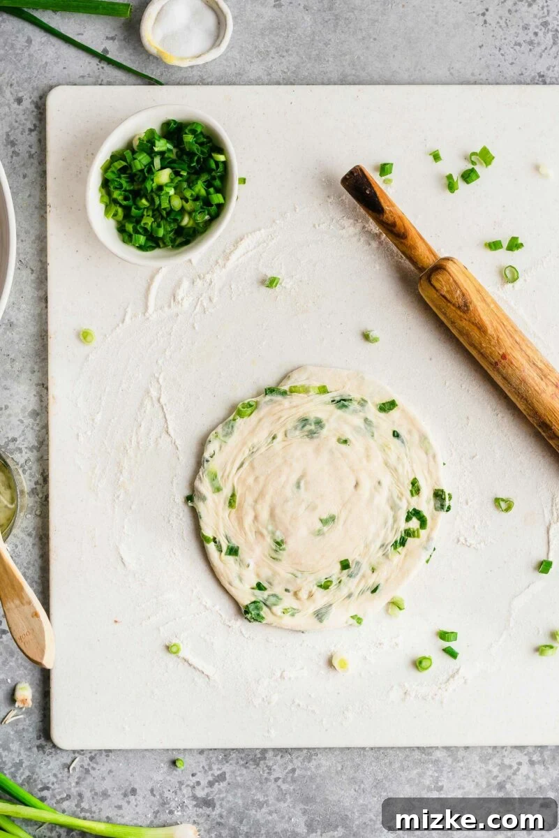 The final roll: flattening the coiled dough into a circle, preserving the internal layers for maximum crispness and chewiness. Overhead view of a coiled scallion pancake dough, flattened and rolled out into a circular shape, ready for cooking.