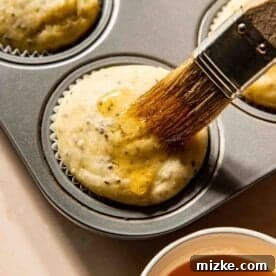 A pastry brush applies melted honey to freshly baked muffins in a muffin tin, with a small cup of honey nearby, showcasing the final touch.