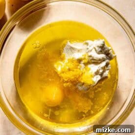 Glass bowl with egg, lemon zest, ricotta, oil, and other liquid ingredients. Halved lemons are beside the bowl on a marble surface, ready to be mixed.