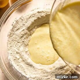 Wet batter is being poured into a well in a bowl of dry flour mixture, with two halved lemons nearby, illustrating the mixing process.