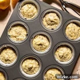 A muffin tray with twelve paper liners filled with uncooked lemon chia seed batter, ready for baking. A lemon and a scoop with batter are nearby on the countertop.