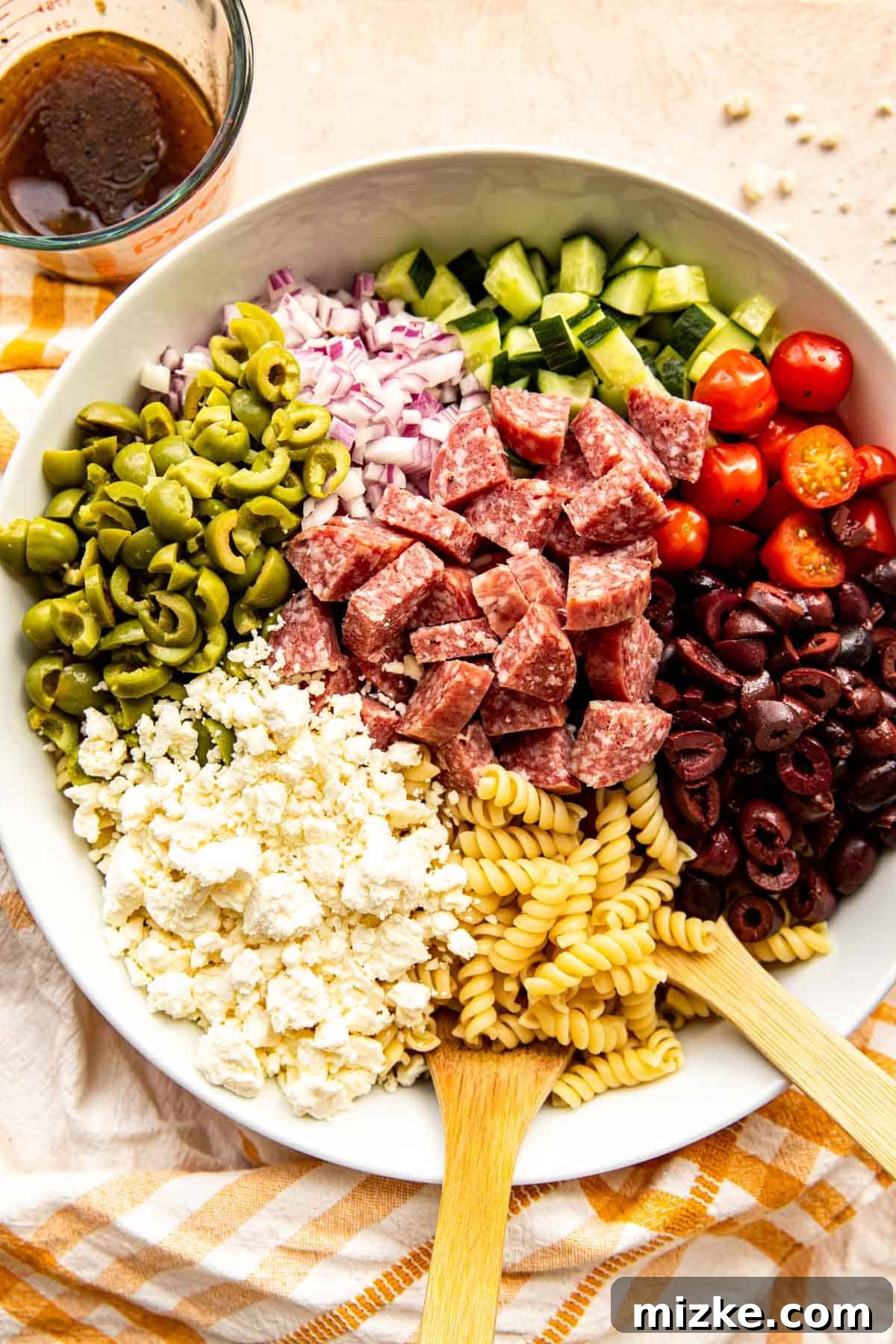 Pasta Salad Ingredients Assembled A large bowl filled with cooked pasta, colorful olives, red onions, tomatoes, salami, and crumbled feta cheese, with a measuring cup of dressing ready on the side.