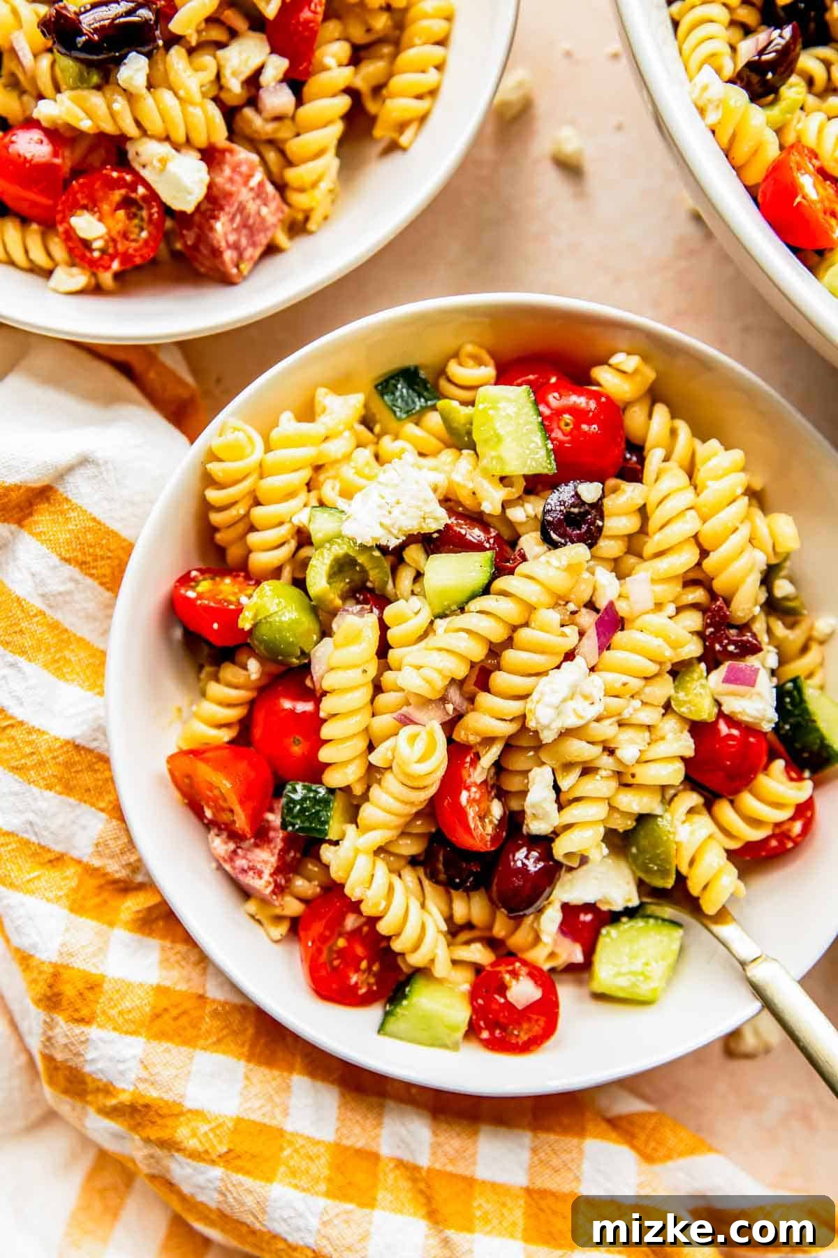 Chilled Mediterranean Pasta Salad A perfectly chilled Mediterranean pasta salad served in a elegant white bowl, placed next to a charming gingham yellow towel, ready to be enjoyed.