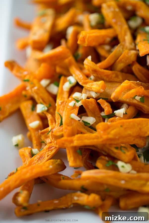 These baked garlic sweet potato fries are a great side dish for any burger or whenever the craving hits! Recipe on tablefortwoblog.com Close-up of golden baked sweet potato fries seasoned with fresh garlic and herbs, highlighting their crispy texture. Recipe on tablefortwoblog.com