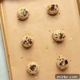 Six perfectly portioned cookie dough balls, studded with chocolate and pistachios, arranged on a parchment-lined baking sheet, ready for the oven.