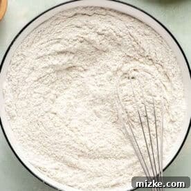 A metal whisk blending all-purpose flour, bread flour, cornstarch, baking soda, salt, and coffee grounds in a white bowl, ensuring a uniform dry mix.
