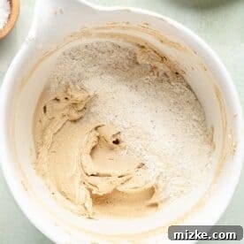 The dry flour mixture is gradually being added to the butter and sugar blend in a white stand mixer bowl, being mixed on low speed.