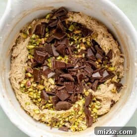 Chopped dark chocolate chunks and pistachios are being gently folded into the cookie batter in a white stand mixer bowl with a spatula.