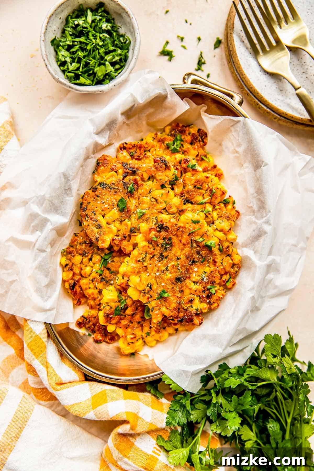 Smoky Sweet Corn Bites 2 Golden brown corn fritters arranged neatly on parchment paper in a metal container, garnished with fresh parsley, beside a charming gingham linen towel.