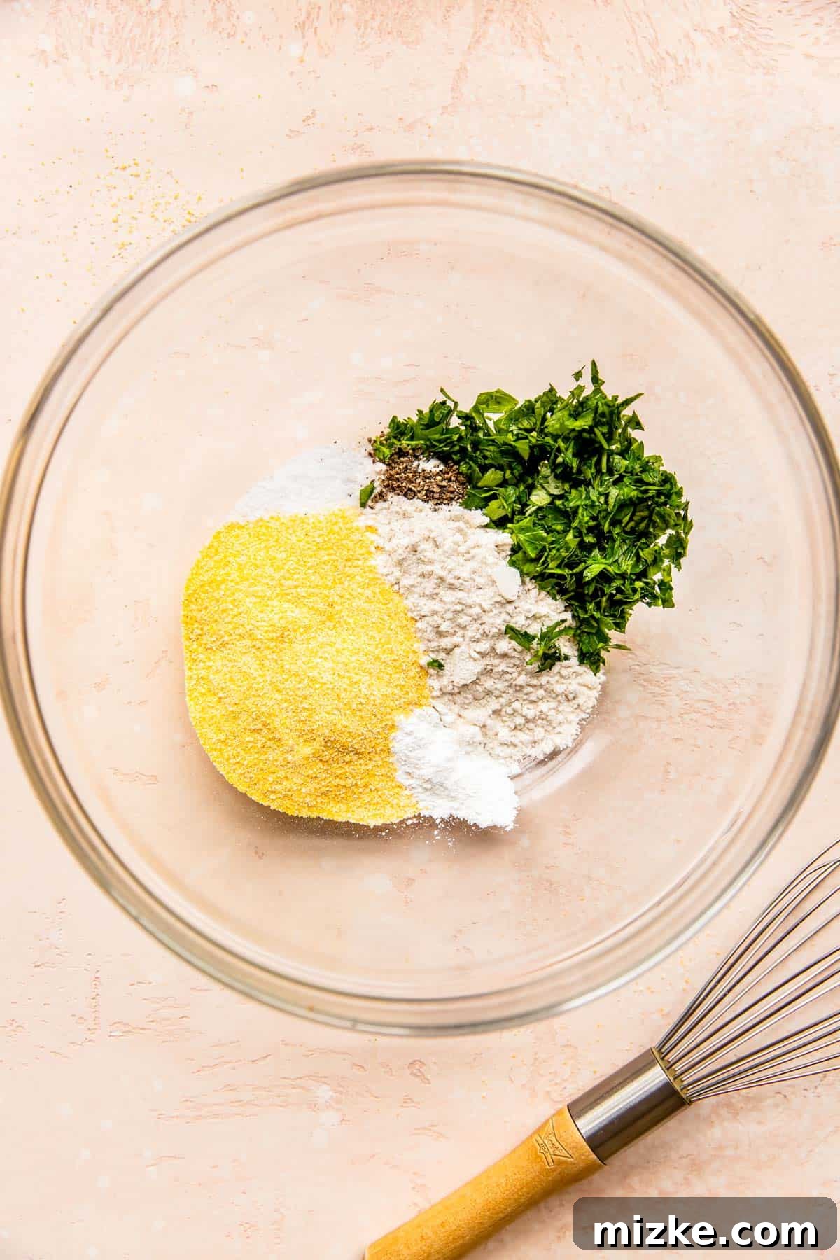 Smoky Sweet Corn Bites 3 A clear bowl holds the dry ingredients for corn fritters: cornmeal, flour, fresh parsley, baking powder, salt, and black pepper, next to a metal whisk.