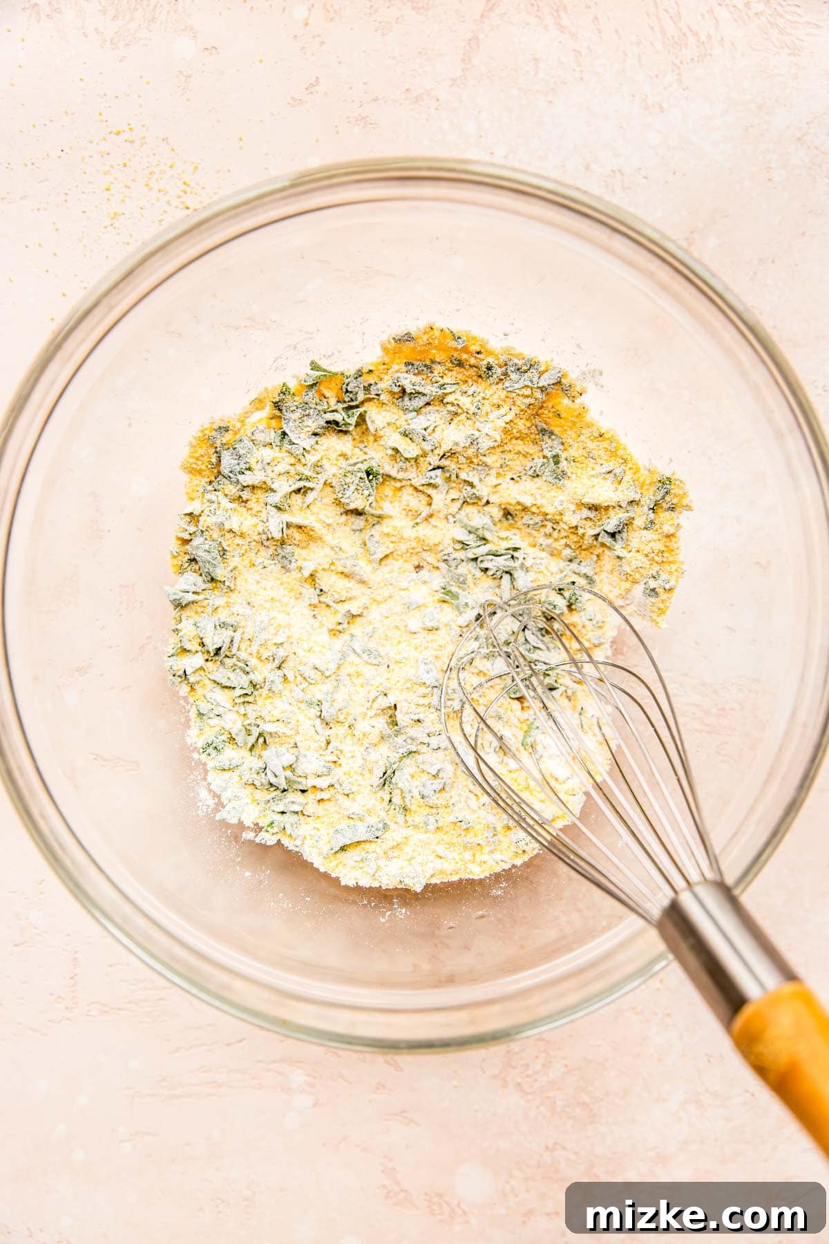 Smoky Sweet Corn Bites 4 A metal whisk is used to thoroughly combine cornmeal, flour, fresh parsley, baking powder, salt, and pepper in a clear mixing bowl, forming the base of the fritter batter.
