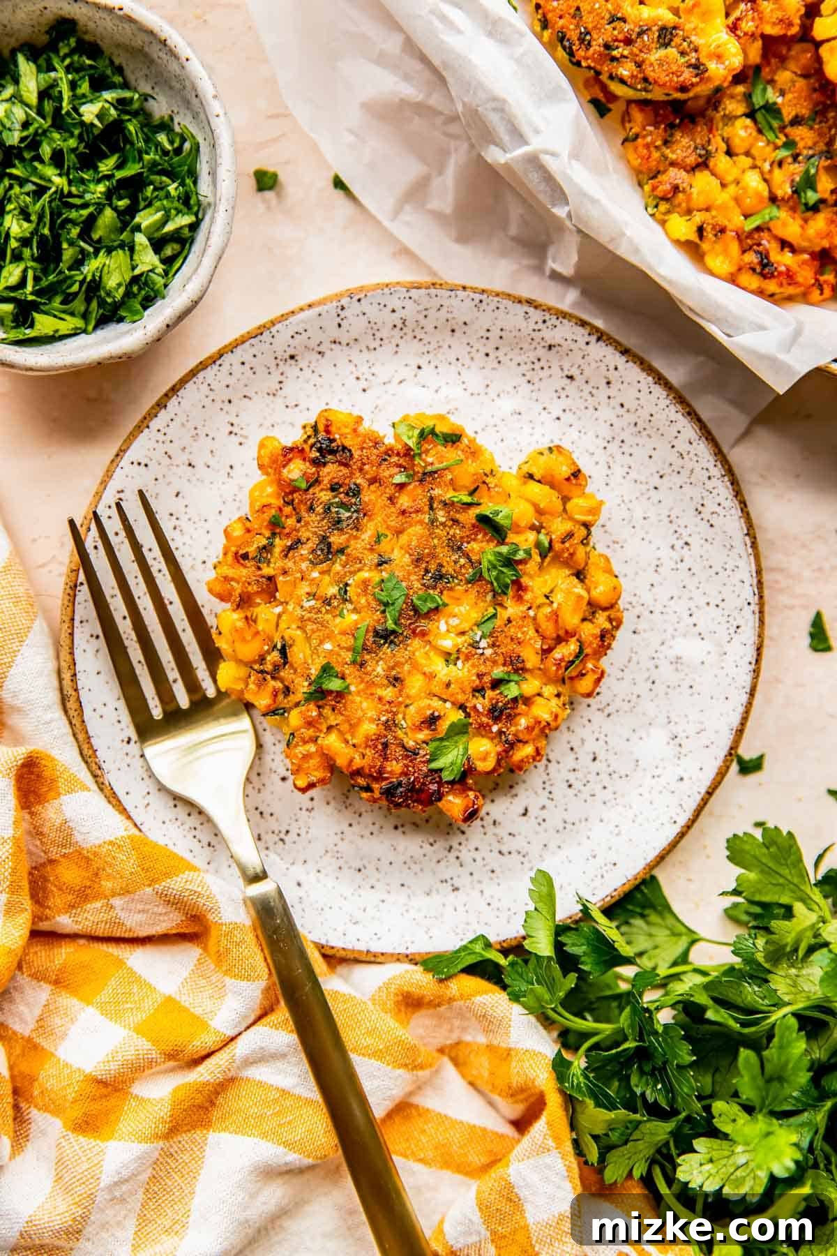 Smoky Sweet Corn Bites 8 A single, perfectly golden corn fritter sits on a speckled ceramic plate, accompanied by an elegant gold metal fork, ready to be tasted.