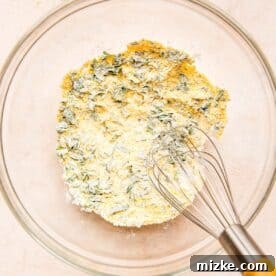 A metal whisk in a clear bowl mixing together cornmeal, flour, fresh parsley, baking powder, salt, and pepper.