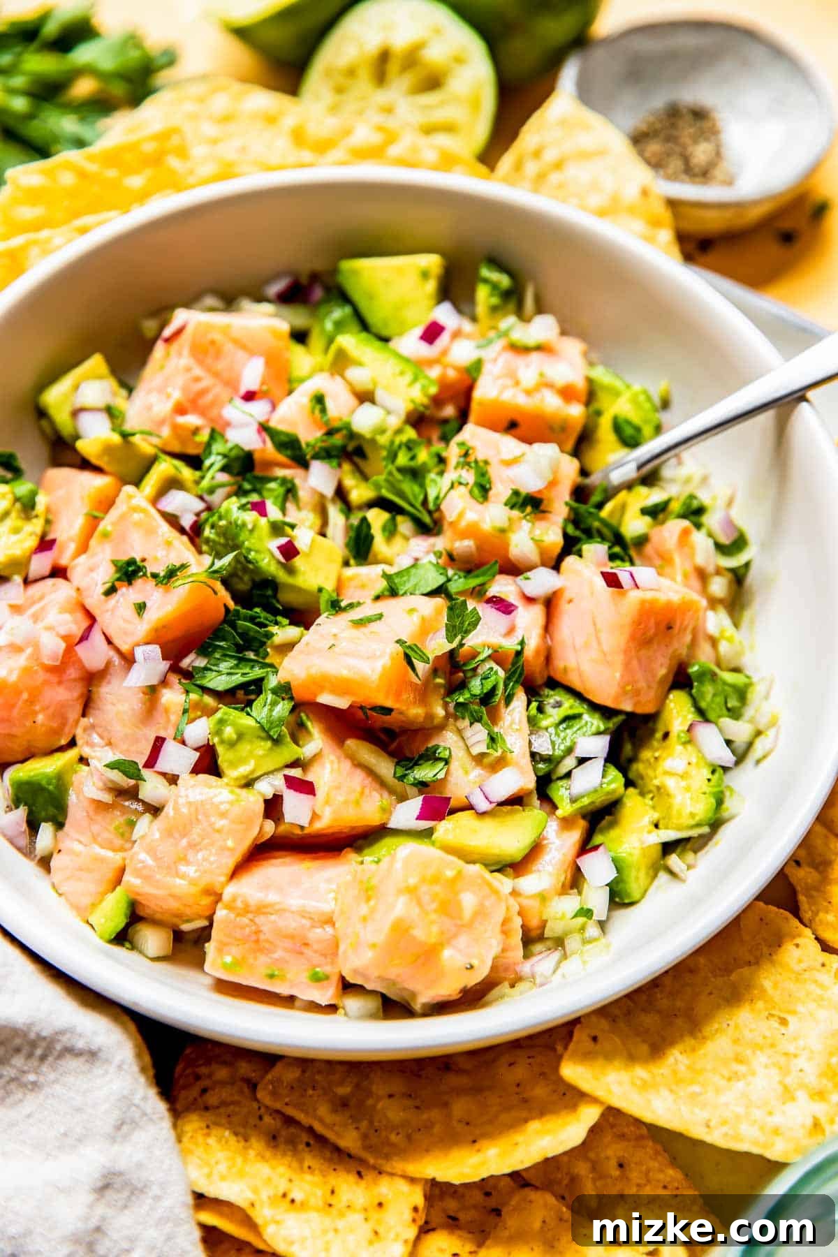 Zesty Salmon Ceviche 3 A serving of salmon ceviche in a white bowl, accompanied by a metal spoon and a scattering of golden tortilla chips.