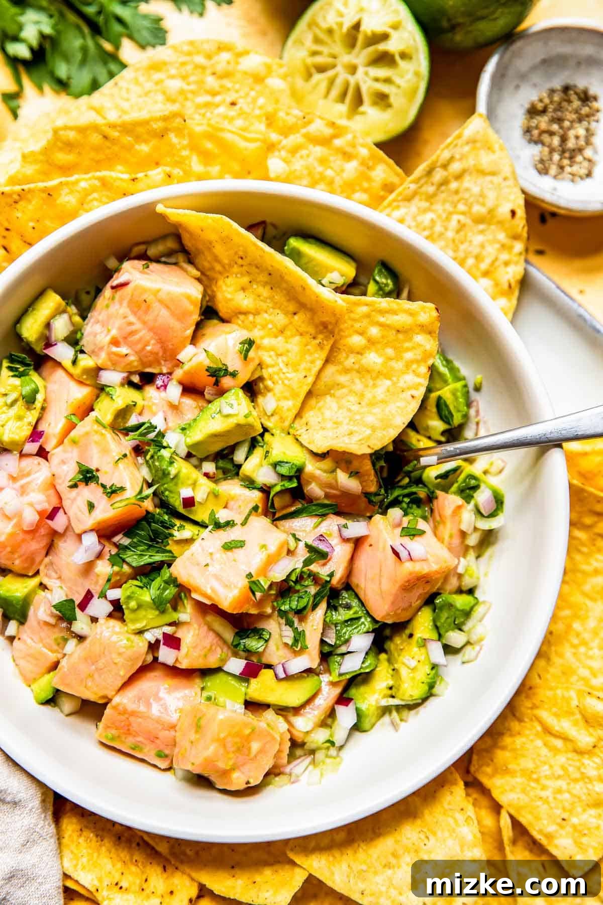 Zesty Salmon Ceviche 4 An overhead view of salmon ceviche served in a white bowl, with a metal spoon nestled within the mixture and two tortilla chips peeking out.
