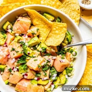 An overhead image of salmon ceviche in a white bowl with a metal spoon and two tortilla chips in the bowl, ready to be served.