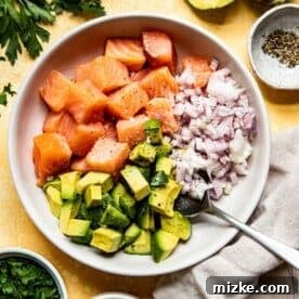 Cubed salmon, avocado, and shallots tossed together with lime juice in a bowl, alongside freshly juiced limes.