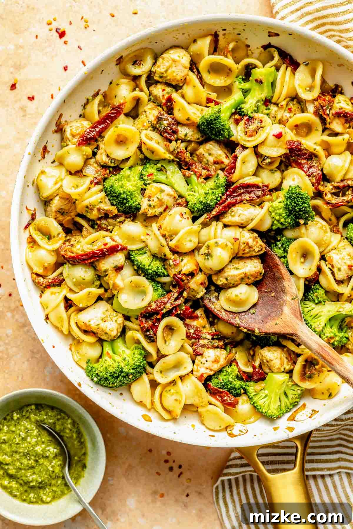 Vibrant Basil Pesto Chicken Pasta 2 A skillet filled with orecchiette pasta, bright green broccoli florets, rich red sun-dried tomatoes, and savory chicken pieces. A wooden spoon rests within the skillet, inviting a taste. A small bowl of vibrant green basil pesto is placed thoughtfully next to the skillet on a rustic surface.