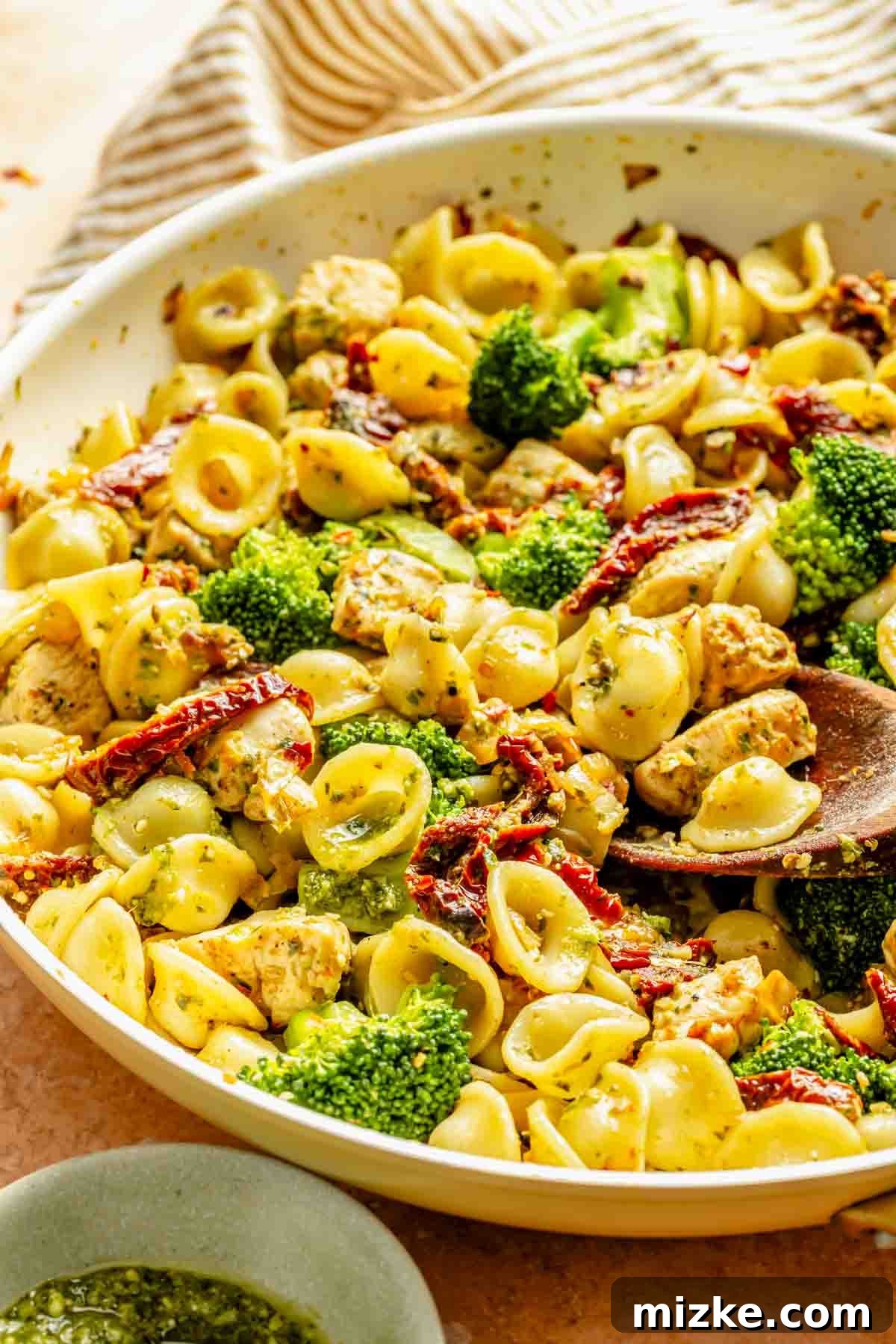 Vibrant Basil Pesto Chicken Pasta 3 A beautifully plated bowl of orecchiette pasta, generously mixed with perfectly cooked chicken pieces, vibrant green broccoli florets, and chewy sun-dried tomatoes. The dish is lusciously tossed in a rich basil pesto sauce. A wooden spoon is partially visible, inviting you to dig in.