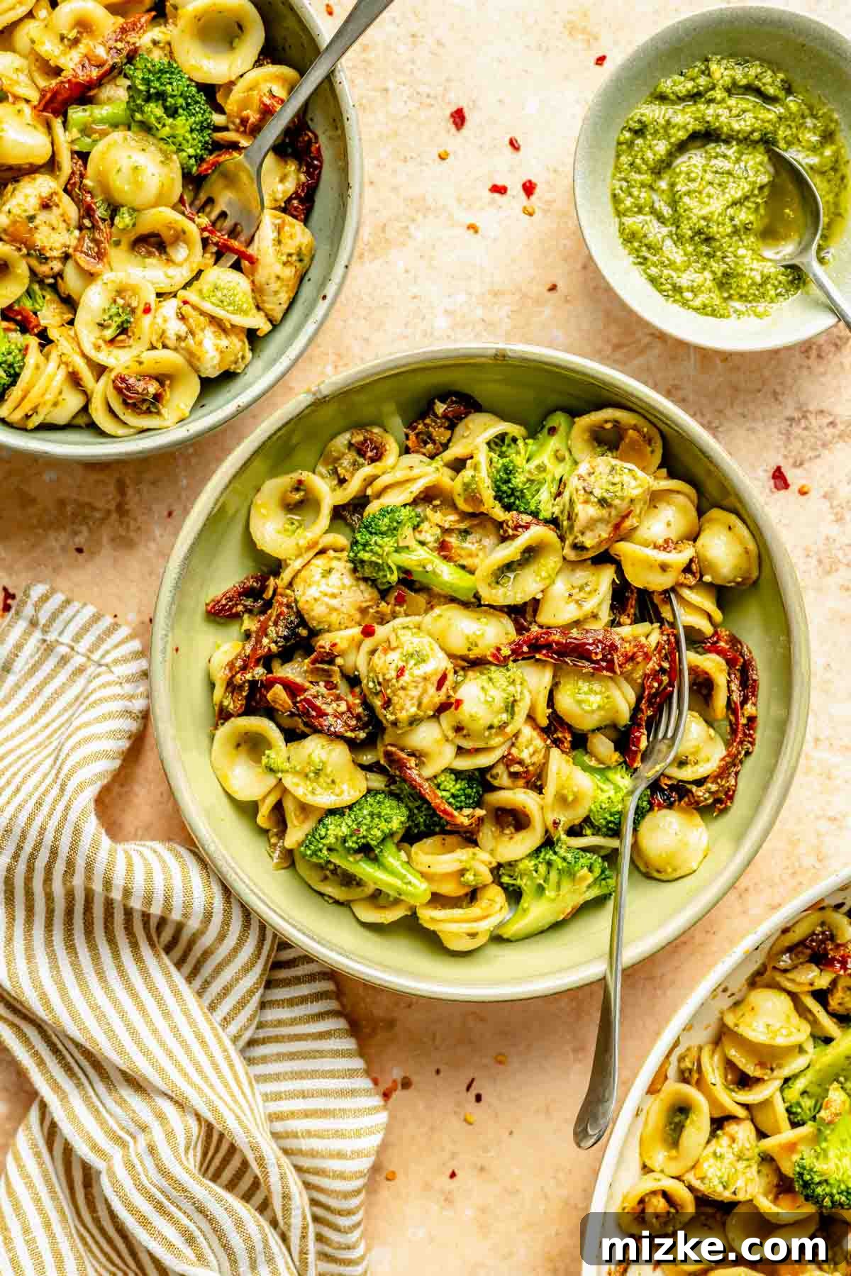 Vibrant Basil Pesto Chicken Pasta 4 Two serving bowls of fresh orecchiette pasta, vibrant with broccoli, sun-dried tomatoes, and chicken, generously coated in pesto. A striped linen napkin and a small bowl of extra basil pesto are stylishly arranged beside them, ready to be enjoyed.