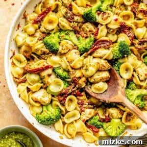 A skillet filled with orecchiette pasta, broccoli, sun-dried tomatoes, and chicken pieces. A wooden spoon is in the skillet. A small bowl of basil pesto is next to the skillet.