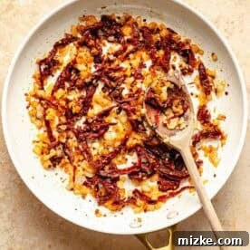 A white frying pan with sautéed onions and sun-dried tomatoes, and a wooden spoon on a beige surface.