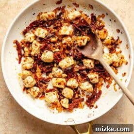 A skillet with cooked chicken, sun-dried tomatoes, and onions, stirred with a wooden spoon on a beige surface.
