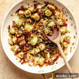 A pan filled with cooked chicken, mixed with sun-dried tomatoes, spices, and pesto, is being stirred with a wooden spoon on a beige background.