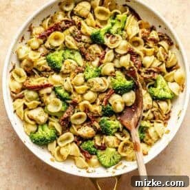 A skillet of orecchiette pasta with pieces of chicken, broccoli, and sun-dried tomatoes, stirred with a wooden spoon.