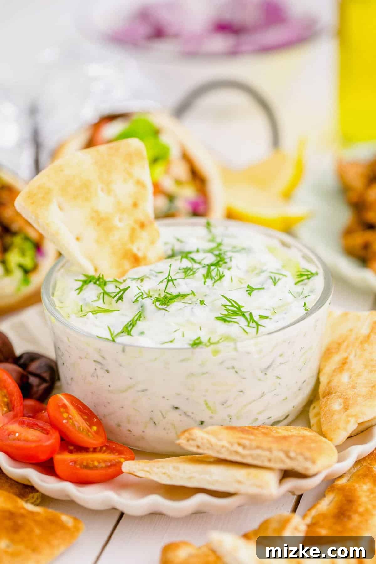 Creamy Homemade Tzatziki 2 Creamy Tzatziki dip served in a clear glass bowl, artfully arranged on a plate with golden pita chips, ripe olives, and halved grape tomatoes, inviting a delicious snack experience.