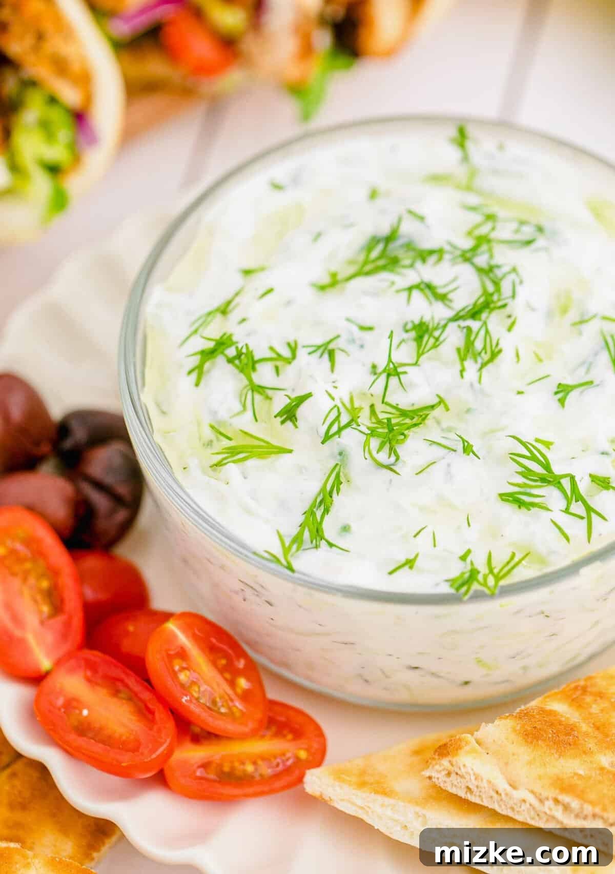 Creamy Homemade Tzatziki 3 A vibrant close-up of tzatziki dip nestled in a glass bowl, beautifully complemented by fresh olives, ripe grape tomatoes, and soft pita bread, creating an inviting Mediterranean spread.
